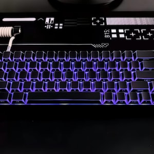 May include: A black mechanical keyboard with purple LED backlighting. The keyboard has a compact layout with a white coiled cable. The keyboard is on a black surface with white graphic designs.