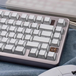 May include: A white and silver mechanical keyboard with white keycaps. The keyboard has a compact layout with function keys and arrow keys. The keys are labeled with white lettering. The keyboard is on a blue surface.