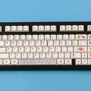 May include: A black mechanical keyboard with white keycaps. The keycaps feature colorful illustrations and symbols. The keyboard sits on a light blue surface. The keyboard has a standard QWERTY layout.
