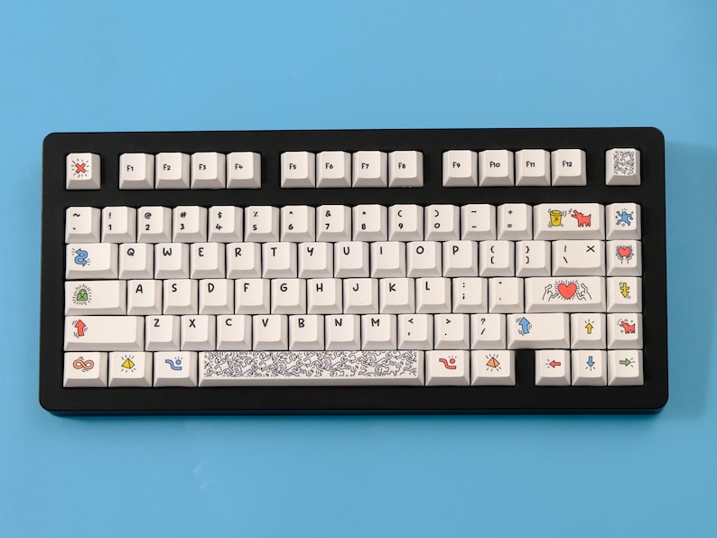 May include: A black mechanical keyboard with white keycaps. The keycaps feature colorful illustrations and symbols. The keyboard sits on a light blue surface. The keyboard has a standard QWERTY layout.