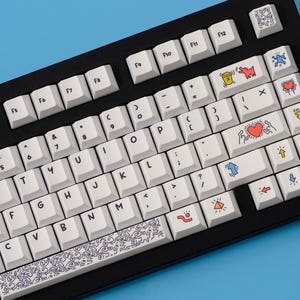 May include: Black mechanical keyboard with white keycaps. Some keycaps feature colorful Keith Haring-style art. The keyboard sits against a blue background. The keycaps have a variety of symbols and letters.