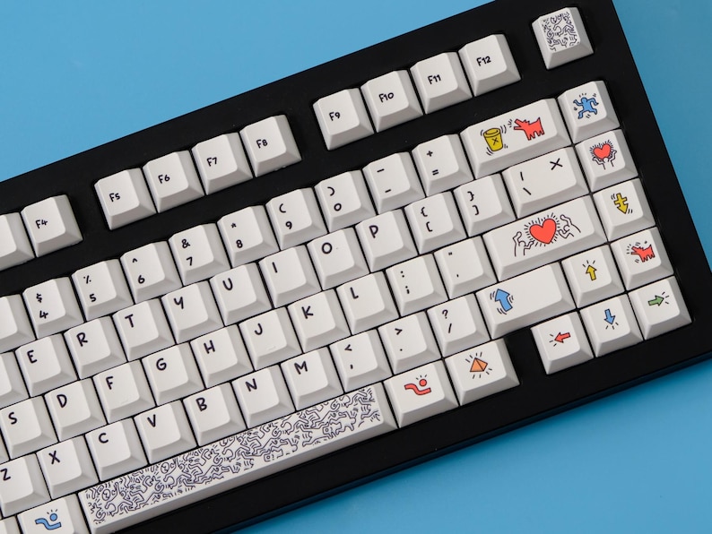 May include: Black mechanical keyboard with white keycaps. Some keycaps feature colorful Keith Haring-style art. The keyboard sits against a blue background. The keycaps have a variety of symbols and letters.