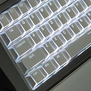 Op de afbeelding: Close-up van een verlicht computertoetsenbord. De toetsen zijn lichtgrijs met witte letters en symbolen. Het toetsenbord heeft een donkergrijze lijst. De toetsen zijn gerangschikt in een standaard QWERTY-indeling, met toetsen als "Esc", "Shift" en "Ctrl" zichtbaar.