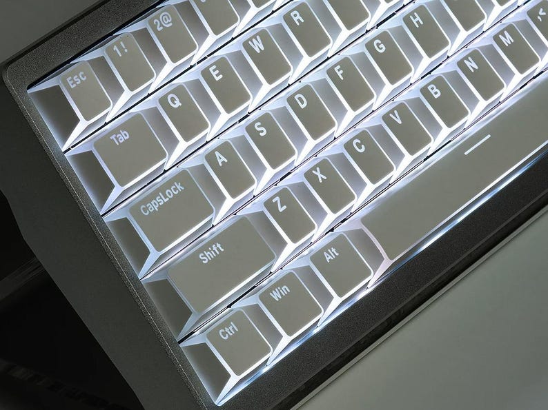 Op de afbeelding: Close-up van een verlicht computertoetsenbord. De toetsen zijn lichtgrijs met witte letters en symbolen. Het toetsenbord heeft een donkergrijze lijst. De toetsen zijn gerangschikt in een standaard QWERTY-indeling, met toetsen als "Esc", "Shift" en "Ctrl" zichtbaar.