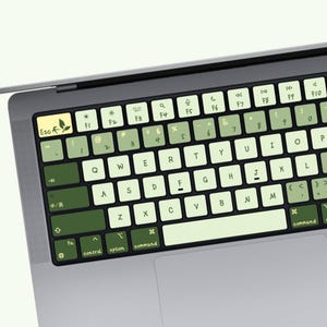 Puede incluir: Una funda para teclado de portátil con un degradado de tonos verdes, desde el verde menta claro hasta el verde bosque profundo. Las teclas muestran diseños QWERTY estándar con letras negras, incluyendo un icono de hoja en la tecla escape.