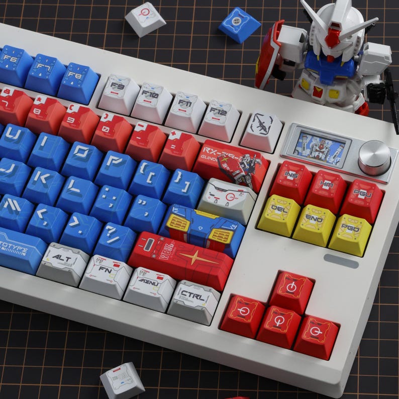 Puede incluir: Un teclado mec&aacute;nico blanco con teclas azules, rojas y blancas. Las teclas tienen varios s&iacute;mbolos y texto, incluyendo "CTRL", "FN" y "ALT". Tambi&eacute;n se ve un peque&ntilde;o modelo Gundam y teclas adicionales.