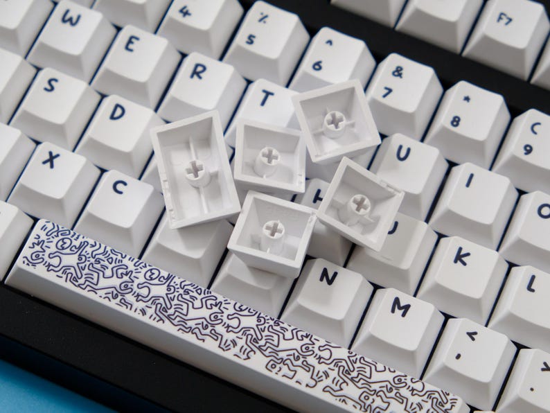May include: Close-up of a white mechanical keyboard with keycaps removed. The keyboard has white keycaps with black lettering. A spacebar with a black and white graphic design is visible.
