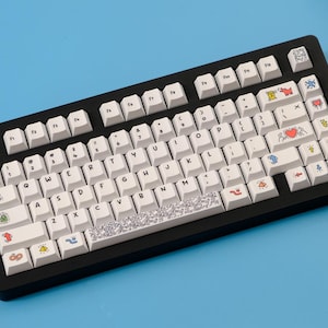 May include: A black mechanical keyboard with white keycaps. The keycaps feature various colorful illustrations, including hearts, butterflies, and abstract designs. The keyboard sits on a blue surface.