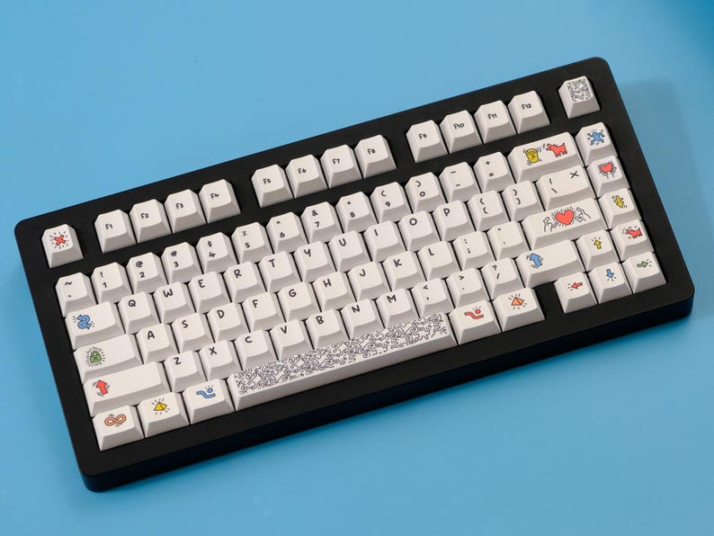 May include: A black mechanical keyboard with white keycaps. The keycaps feature various colorful illustrations, including hearts, butterflies, and abstract designs. The keyboard sits on a blue surface.