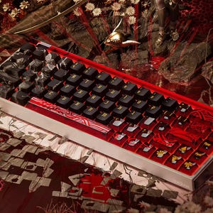 Może przedstawiać: Niestandardowa klawiatura mechaniczna z czarnymi nakładkami na klawisze i złotymi literami. Klawiatura ma czerwoną i białą ramkę ze skomplikowanymi wzorami. Ogólna estetyka jest ciemna i szczegółowa, z naciskiem na kunszt i design.