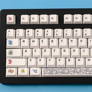 May include: Black mechanical keyboard with white keycaps. Some keycaps feature colorful cartoon graphics and symbols. The keyboard has a standard QWERTY layout with function keys.