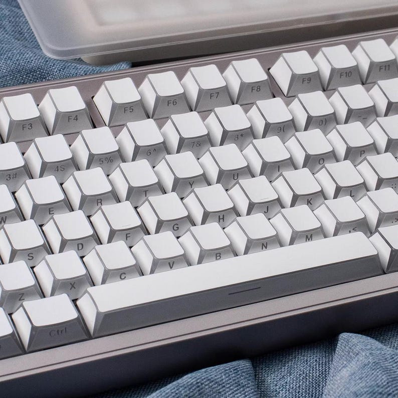 May include: A close-up of a mechanical keyboard with white keycaps and a gray frame. The keys are arranged in a standard QWERTY layout, with function keys along the top. The keyboard is on a denim surface.