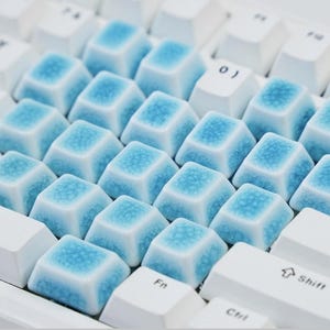 以下が含まれることがあります： 中央にカスタムキーキャップのグループを備えたメカニカルキーボード。これらのキーキャップは白いベースで、上面には質感のあるひび割れた青い釉薬仕上げが施されており、周囲の標準的な白いキーと対照的です。