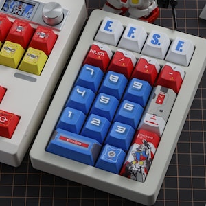 Puede incluir: Un teclado mec&aacute;nico personalizado con teclas blancas, rojas, azules y amarillas. Las teclas presentan varios s&iacute;mbolos, n&uacute;meros y texto, incluyendo "E.F.S.E" y "NUM". El teclado tiene un marco blanco y una peque&ntilde;a pantalla.