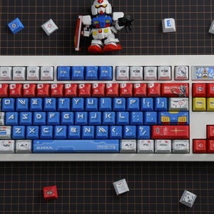 Puede incluir: Un teclado mec&aacute;nico blanco con teclas rojas, azules y blancas. Las teclas presentan varios gr&aacute;ficos y texto, incluida la palabra "GUNDAM". Una peque&ntilde;a figura de Gundam y teclas adicionales est&aacute;n esparcidas alrededor del teclado.