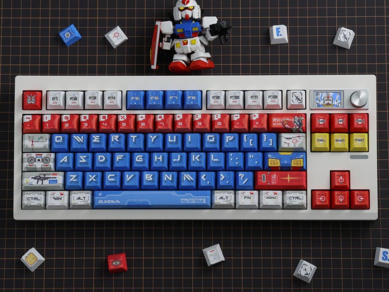 Puede incluir: Un teclado mec&aacute;nico blanco con teclas rojas, azules y blancas. Las teclas presentan varios gr&aacute;ficos y texto, incluida la palabra "GUNDAM". Una peque&ntilde;a figura de Gundam y teclas adicionales est&aacute;n esparcidas alrededor del teclado.