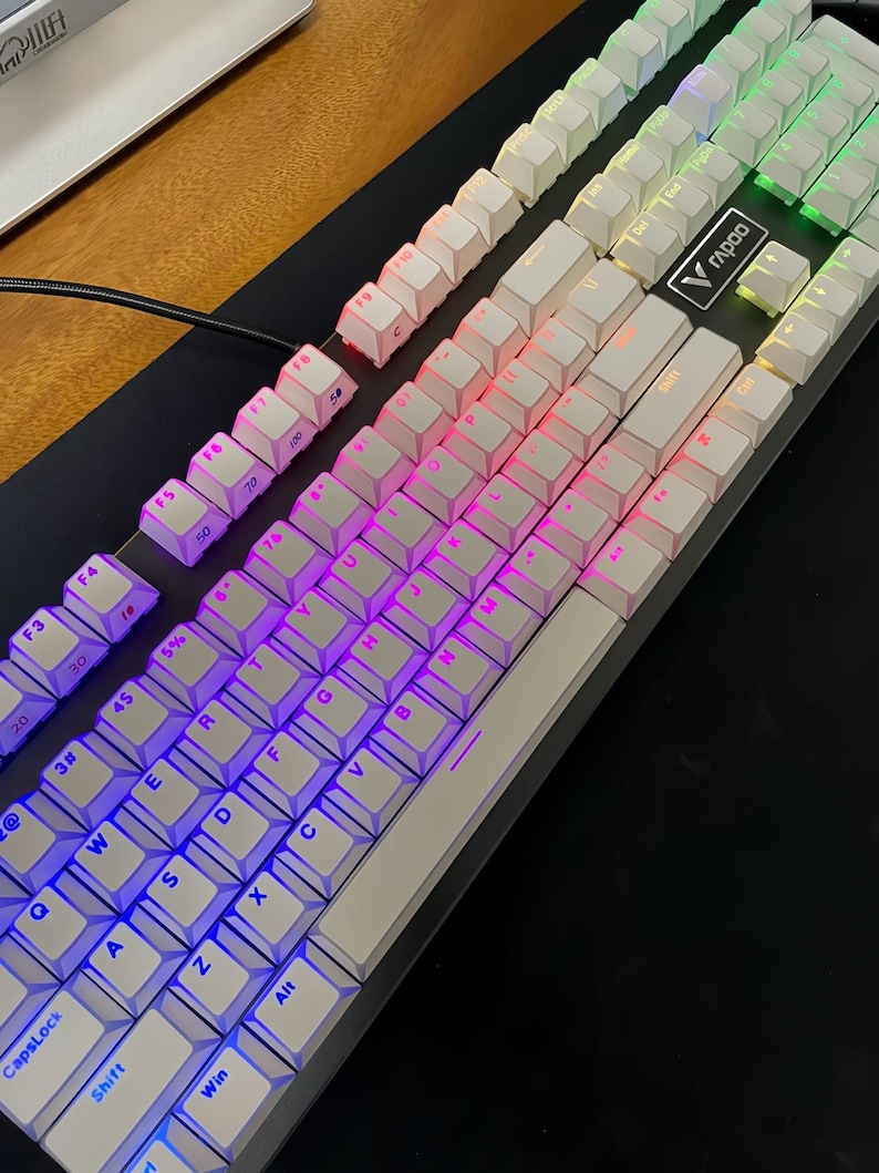 Op de afbeelding: Een witte mechanische toetsenbord met verlichte toetsen. De toetsen zijn verlicht met een kleurengradi&euml;nt van blauw, paars, roze en groen. Het toetsenbord heeft een zwarte basis en een zilveren frame. De merknaam "Rapoo" is zichtbaar.