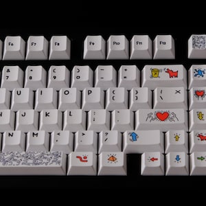 May include: White mechanical keyboard with custom keycaps featuring colorful Keith Haring-style artwork. The keycaps display various figures, hearts, and symbols in red, blue, yellow, and black. The keyboard has a black background.