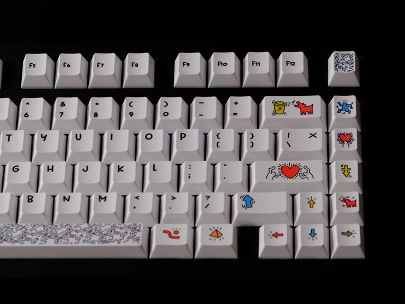 May include: White mechanical keyboard with custom keycaps featuring colorful Keith Haring-style artwork. The keycaps display various figures, hearts, and symbols in red, blue, yellow, and black. The keyboard has a black background.