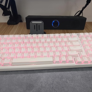 Op de afbeelding: Wit mechanisch toetsenbord met roze LED-achtergrondverlichting. Het toetsenbord ligt op een grijze ondergrond, met een zwarte luidspreker en een telefoonstandaard op de achtergrond. De toetsen zijn wit met roze verlichting.