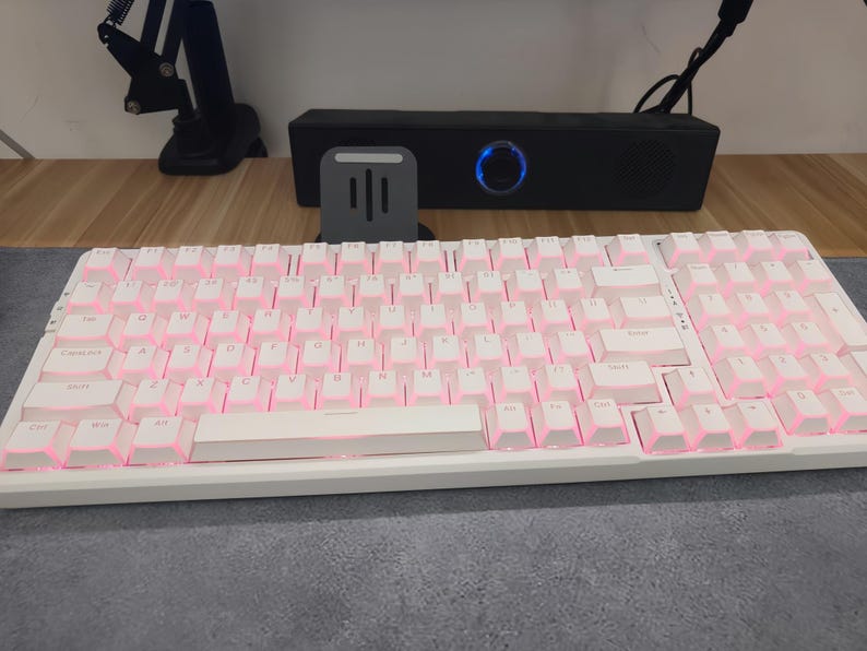 Op de afbeelding: Wit mechanisch toetsenbord met roze LED-achtergrondverlichting. Het toetsenbord ligt op een grijze ondergrond, met een zwarte luidspreker en een telefoonstandaard op de achtergrond. De toetsen zijn wit met roze verlichting.