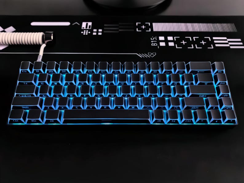 May include: A black mechanical keyboard with blue LED backlighting. The keyboard has a compact design with a 60% layout. The keys are black with white lettering. The keyboard is on a black surface with white graphic designs.