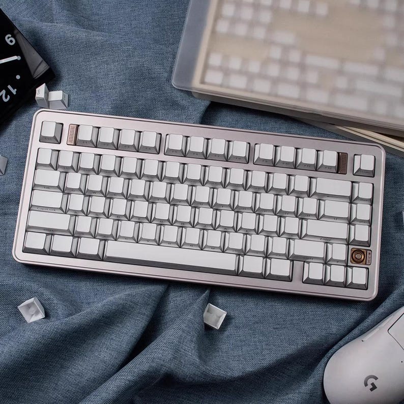 May include: A silver and white mechanical keyboard with a rose gold frame. The keyboard has white keycaps and a few brown accent keys. A white mouse and extra keycaps are also visible.