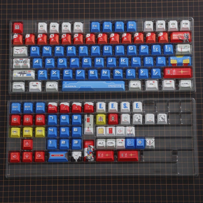 Puede incluir: Un juego de teclas de teclado mec&aacute;nico personalizadas en una bandeja de pl&aacute;stico transparente. Las teclas son principalmente rojas, blancas y azules, con algunos acentos amarillos. Las teclas presentan varios s&iacute;mbolos y texto, incluyendo "GUNDAM" y "PHOTOTYPE".