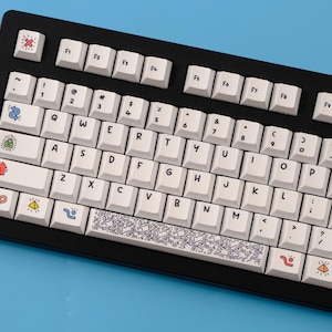 May include: Black mechanical keyboard with white keycaps. The keycaps feature colorful icons and a Keith Haring-style design on the spacebar. The keyboard has a black frame and is set against a blue background.