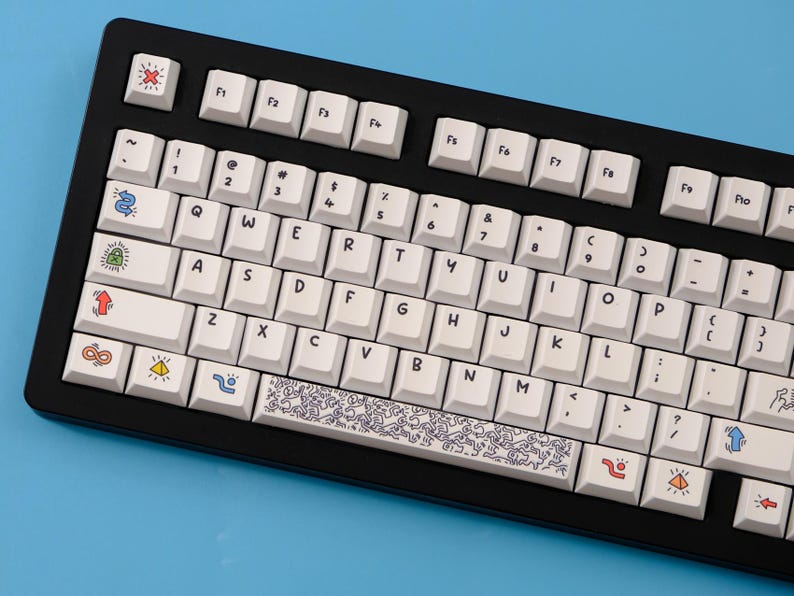 May include: Black mechanical keyboard with white keycaps. The keycaps feature colorful icons and a Keith Haring-style design on the spacebar. The keyboard has a black frame and is set against a blue background.