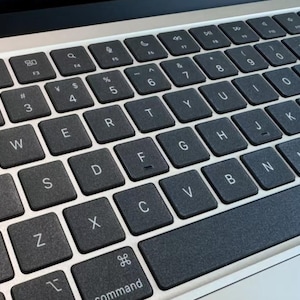 May include: A laptop keyboard featuring dark gray square keys with white lettering in a QWERTY layout. The keys are set against a light silver metallic base, including a command key and a row of function keys along the top edge of the device.