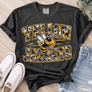 Puede incluir: Una camiseta de manga corta gris carbón con el texto YELLOW JACKETS en fuente de bloque con un patrón de camuflaje amarillo, negro y blanco. Una abeja de dibujos animados con rayas amarillas y negras está centrada entre las palabras.