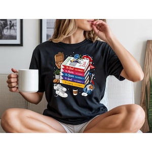 Peut inclure: T-shirt noir avec une illustration de livres empilés intitulés "The Deal", "The Mistake", "The Score", "The Goal" et "The Legacy". Le motif comprend également une boîte à pizza, des baskets, des cookies et un casque.
