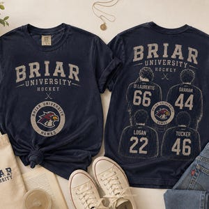 Puede incluir: Una camiseta azul marino con gráficos de hockey de la Universidad Briar. El frente muestra el nombre del equipo y un logo de halcón. La parte posterior muestra cuatro jugadores de hockey con los nombres Di Laurentis 66, Graham 44, Logan 22 y Tucker 46.