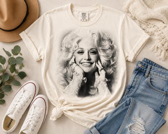 Może przedstawiać: Kremowy t-shirt z krótkim rękawem z czarno-białym nadrukiem fotograficznym uśmiechniętej osoby o bujnych blond włosach, noszącej pierścionki na palcach i odzież z koronkowymi mankietami, z widoczną metką marki Comfort Colors.