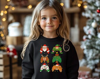 May include: A black sweatshirt featuring six Christmas-themed controller designs. The designs include Santa Claus, an elf, a reindeer, a Christmas tree, a snowman, and a gingerbread man. The sweatshirt is worn by a young girl with blonde hair.