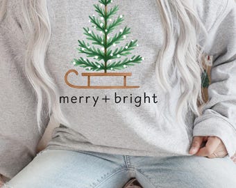 May include: A light gray sweatshirt featuring a green Christmas tree design on a brown sled, with the text "merry + bright" in black. The tree has white accents. The sweatshirt is long-sleeved and made of a soft material.
