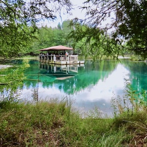 May include: A scenic view of a clear, turquoise-colored lake surrounded by lush green trees and vegetation. A wooden structure with a red roof sits on the water, with people visible on the structure. The sky is overcast.