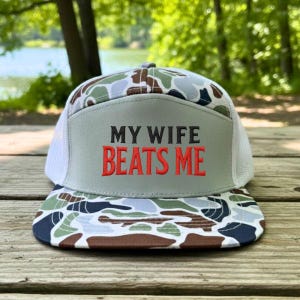 May include: A trucker hat with a camouflage pattern on the brim and top, and a white mesh back. The front panel is gray with the embroidered text "MY WIFE BEATS ME" in red and black.