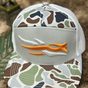 May include: A gray and white trucker hat with a camouflage pattern on the brim and crown. The hat features an embroidered orange and white logo on the front panel. The back of the hat is white mesh.