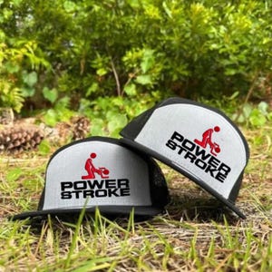 May include: Two black and white trucker hats with the words "POWER STROKE" in black and a red graphic. The hats are resting on green grass with a blurred background of foliage.