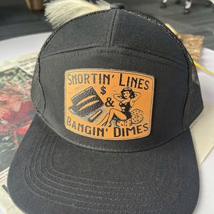 May include: Black trucker hat with a rectangular patch featuring the text "SNORTIN' LINES & BANGIN' DIMES". The patch has a vintage design with a woman, dice, and stacks of money. The hat has a mesh back.