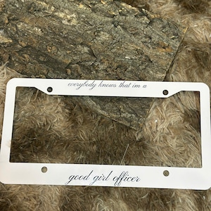 May include: White license plate frame with the text "everybody knows that im a" at the top and "good girl officer" at the bottom. The frame has four mounting holes and is set against a textured background.