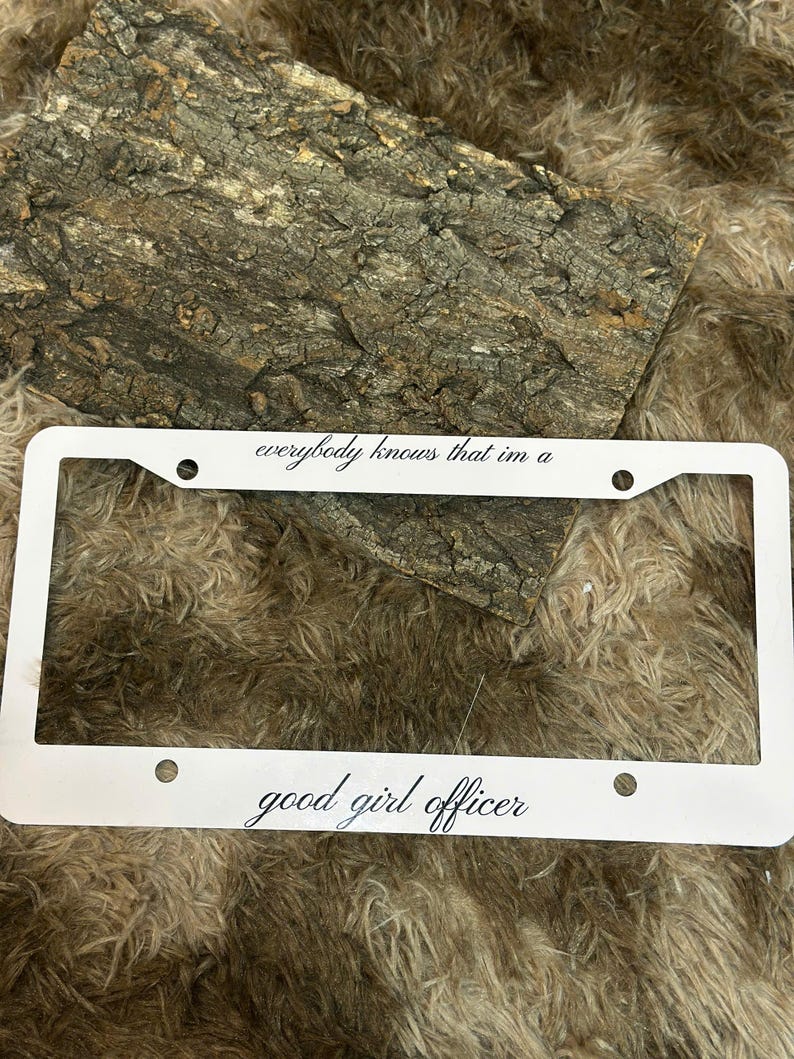 May include: White license plate frame with the text "everybody knows that im a" at the top and "good girl officer" at the bottom. The frame has four mounting holes and is set against a textured background.