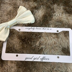 May include: A white license plate frame with the text "everybody knows that im a good girl officer" in a script font. A cream-colored bow tie is in the upper left corner. The background is a brown, textured surface.