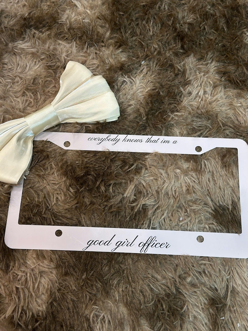 May include: A white license plate frame with the text "everybody knows that im a good girl officer" in a script font. A cream-colored bow tie is in the upper left corner. The background is a brown, textured surface.