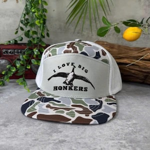 May include: A camouflage trucker hat with a white mesh back and a flat brim. The front panel is light gray with the text "I LOVE BIG HONKERS" and a graphic of a goose in flight. The brim and top of the hat feature a camouflage pattern.
