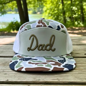 May include: A camouflage print baseball cap with the word "Dad" embroidered in a cursive font on a light gray patch. The cap has a white mesh back and a curved brim.
