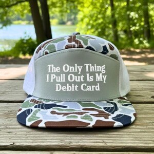 May include: A trucker hat with a camouflage pattern on the bill and top, and a gray front panel. The front panel has the text "The Only Thing I Pull Out Is My Debit Card" in white. The hat has a white mesh back.