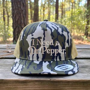 May include: Camouflage trucker hat with the text "I Need a Dr Pepper" embroidered on the front. The hat features a mesh back and a curved brim. The hat is sitting on a wooden surface.